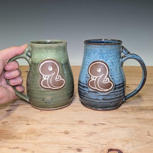 Peut inclure: Deux tasses en céramique avec anses, l'une vert sauge chiné et l'autre bleu et gris foncé nuancé. Chaque tasse présente un emblème central en argile brune en relief représentant une créature arrondie stylisée avec un seul œil visible.