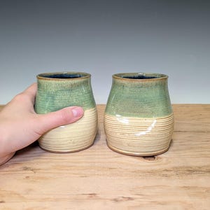 May include: Two ceramic tumblers featuring a two-tone glaze design. The upper portion of each vessel is finished in a smooth, mottled sage green, while the lower section displays a cream-colored base with horizontal ribbed texture detailing.