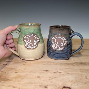 May include: Two ceramic mugs with handles, each featuring a raised brown clay applique of a cartoon elephant. The left mug has a green and cream glaze, while the right mug has a dark blue and brown glaze. Both mugs have a cylindrical shape.