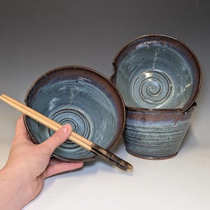May include: Handmade ceramic ramen bowls in shades of blue and brown. The bowls have a spiral design in the center and a textured exterior. A pair of wooden chopsticks rests on one of the bowls. The bowls are ideal for serving soup or noodles.