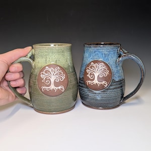 May include: Two ceramic mugs with a tree of life design. One mug is green, the other is blue. Each mug has a brown circular emblem with a white tree design. The mugs have large handles and are suitable for hot beverages.