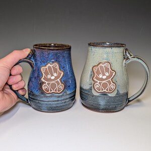 May include: Two handmade ceramic mugs. One is blue with a brown badger applique, the other is light green with a similar badger design. Both mugs have a handle and a dark brown rim. The mugs are approximately 4 inches tall.
