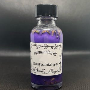 May include: A clear glass bottle with a black cap, filled with purple liquid and dried flowers. The label reads "Commanding Oil" and "edensessential.com" in a decorative font. The bottle is likely an essential oil or potion.