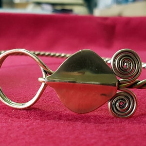 Puede incluir: Una diadema de metal dorado con un diseño de alambre trenzado. La diadema tiene una pieza grande en forma de hoja en el centro y dos diseños en espiral a cada lado.