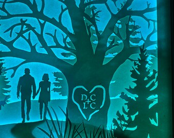 May include: A paper cut-out silhouette of a couple standing in front of a tree with a heart carved into the trunk. The heart has the initials "D+C" and "L+C" inside. The couple is silhouetted against a blue background.
