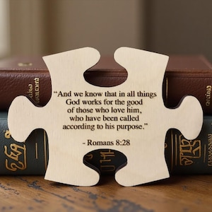 May include: A light-coloured wooden jigsaw piece with a Bible verse engraved on it. The jigsaw is in front of two books with gold lettering. The text on the jigsaw reads, "And we know that in all things God works for the good of those who love him..." Romans 8:28.