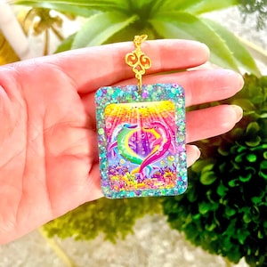 May include: A rectangular pendant with a colorful dolphin design. The pendant features a gold-colored filigree top and a glittery border. The dolphins are in shades of green, pink, and purple, with a yellow and orange background.