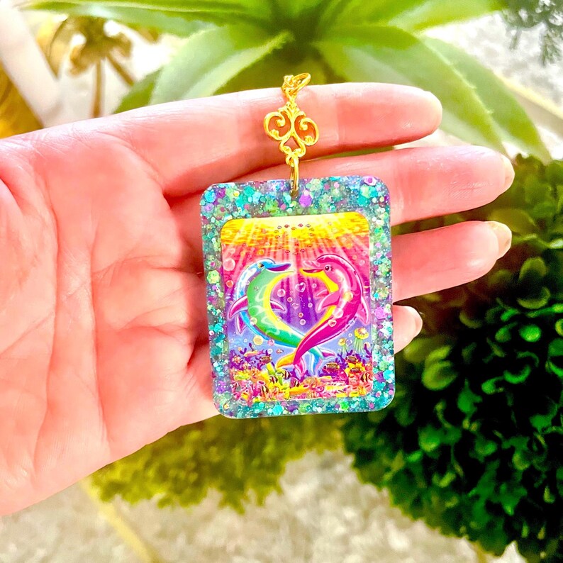 May include: A rectangular pendant with a colorful dolphin design. The pendant features a gold-colored filigree top and a glittery border. The dolphins are in shades of green, pink, and purple, with a yellow and orange background.