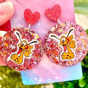 May include: A pair of dangle earrings featuring a pink and gold glitter background with a cartoon image of Pluto the dog. The earrings are topped with a red glitter heart.