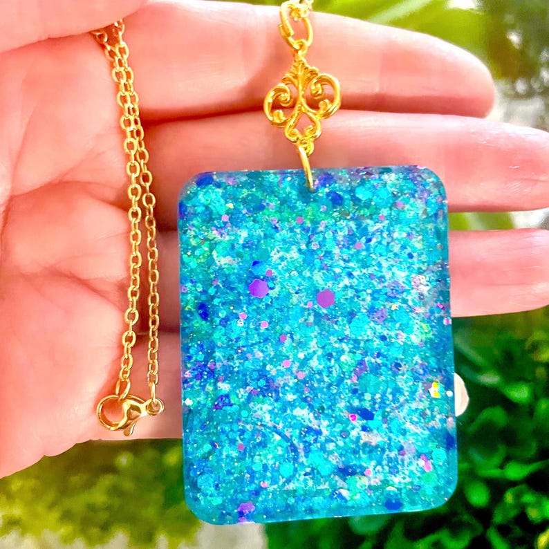 May include: A gold-colored chain necklace with a rectangular pendant. The pendant is filled with blue and purple glitter, creating a sparkling effect. The necklace is held up by a decorative gold-colored bail.