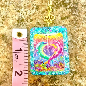 May include: A rectangular pendant necklace featuring a colorful dolphin design. The pendant has a turquoise glitter border and a gold-colored chain. The image inside the pendant shows two dolphins forming a heart shape, with an underwater scene.
