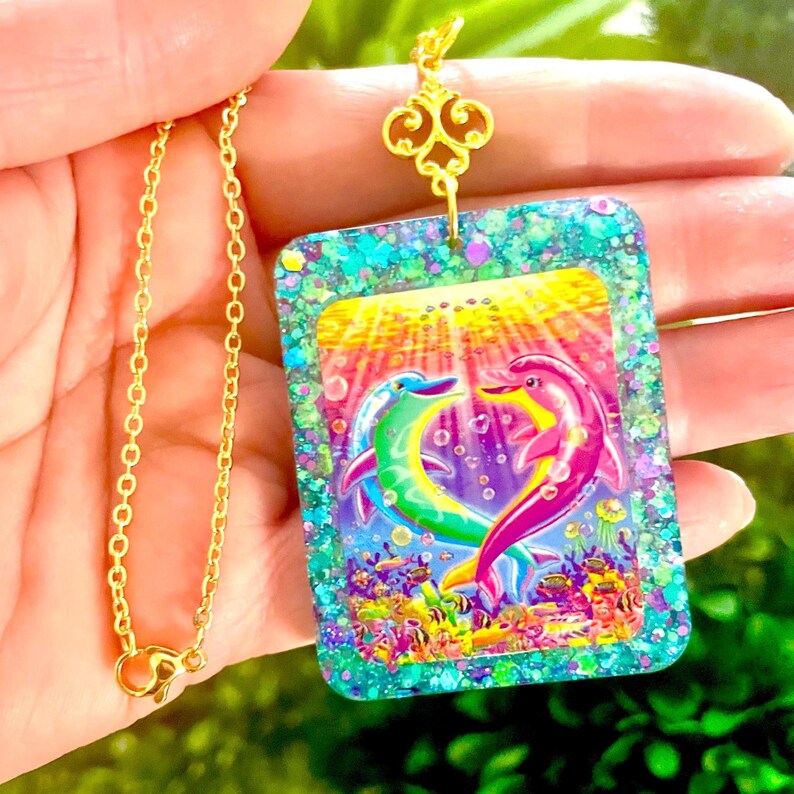 May include: A gold-colored chain necklace with a rectangular pendant featuring two dolphins in a heart shape. The pendant has a colorful underwater scene with glitter accents and a gold-colored decorative top.
