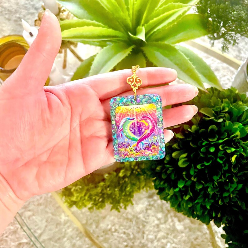 May include: A rectangular pendant with a gold-colored chain. The pendant features a colorful illustration of two dolphins forming a heart shape, set against a vibrant underwater scene. The edges are embellished with glitter.
