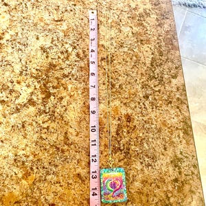 May include: A gold-colored necklace with a pendant featuring a colorful heart design. The pendant is rectangular and measures approximately 1.5 inches in length. The necklace is displayed against a brown surface.