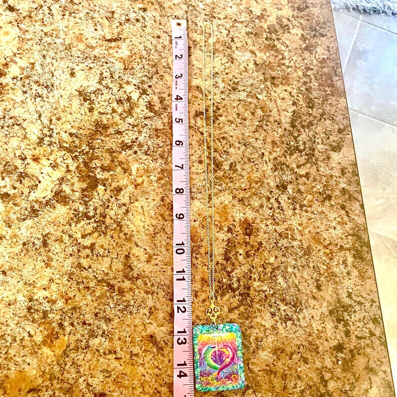 May include: A gold-colored necklace with a pendant featuring a colorful heart design. The pendant is rectangular and measures approximately 1.5 inches in length. The necklace is displayed against a brown surface.