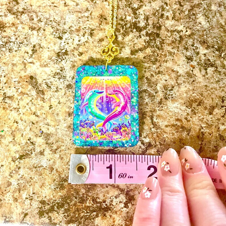 May include: A gold-colored chain necklace with a pendant featuring a colorful dolphin design. The pendant is a square shape with a turquoise glitter border. The dolphins are in shades of green and pink, with a background of an underwater scene.