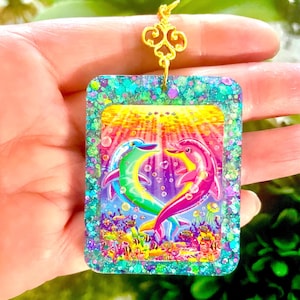 May include: A rectangular pendant with a colorful dolphin design. The pendant features a heart-shaped arrangement of two dolphins in shades of green and pink, set against a vibrant underwater scene. The pendant is framed with glittery teal resin and has a gold-colored decorative bail.