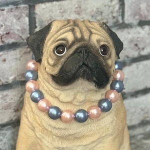 May include: A pug dog figurine wearing a necklace of pink and gray pearls. The figurine is made of a light brown material and has a realistic appearance.