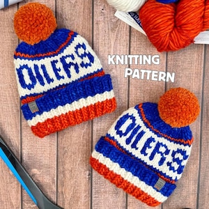 Op de afbeelding: Twee blauwe, witte en oranje gebreide mutsen met het woord "OILERS" in het wit, afgewerkt met oranje pompons. Een hockeystick en een puck zijn ook zichtbaar. De tekst "KNITTING PATTERN" staat in het midden van de afbeelding.