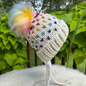 以下が含まれることがあります： クリーム色のニット帽。カラフルなポンポンとマルチカラーのハート型アクセントが特徴です。帽子には編み込みの紐が付いており、木製のスタンドに飾られています。ポンポンは虹色で、冬のアクセサリーに遊び心を加えています。