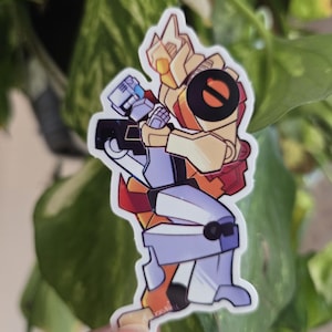 May include: A sticker featuring two cartoon robots embracing. One robot is primarily yellow and orange, with a black circle on its chest. The other robot is blue and white. The sticker is held in front of a background of green leaves.