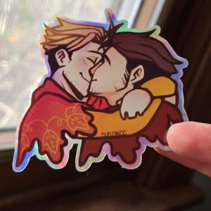 May include: Holographic sticker featuring two cartoon men embracing and kissing. One man has blonde hair and wears a red hooded cloak with leaf designs. The other has brown hair and wears a yellow shirt. The sticker has a shiny, iridescent finish.