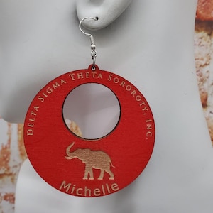 May include: Red circular wooden earrings with a cutout in the center. The earrings feature the text "Delta Sigma Theta Sorority, Inc." and an elephant silhouette. The name "Michelle" is engraved below the elephant.