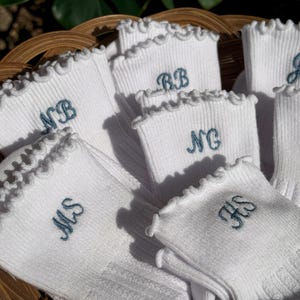 May include: White socks with a ruffled edge and embroidered initials in teal. The socks are arranged in a woven basket. The initials include "MS", "NB", "NG", "RB", and "HS".