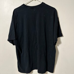 May include: A black t-shirt hanging on a white plastic hanger. The t-shirt is plain and appears to be made of a soft fabric. The shirt is hanging against a white wall.