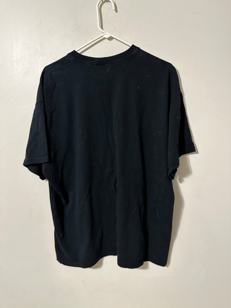 May include: A black t-shirt hanging on a white plastic hanger. The t-shirt is plain and appears to be made of a soft fabric. The shirt is hanging against a white wall.