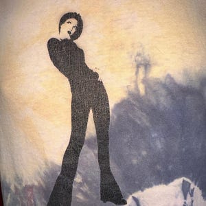 K&ouml;nnte beinhalten: Ein Batik-Shirt mit einer schwarzen Silhouette einer Frau in einem lang&auml;rmeligen Outfit und einer Schlaghose.