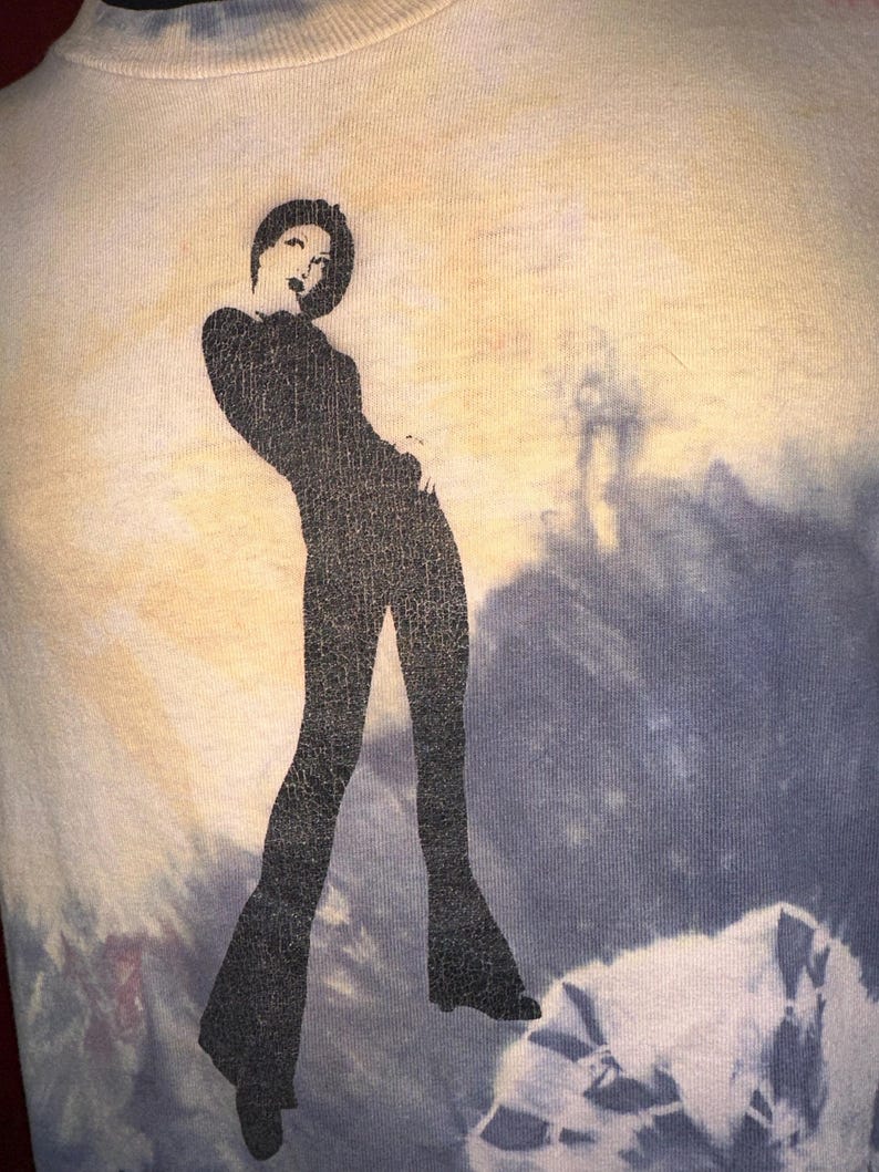 K&ouml;nnte beinhalten: Ein Batik-Shirt mit einer schwarzen Silhouette einer Frau in einem lang&auml;rmeligen Outfit und einer Schlaghose.