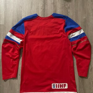 May include: Red hockey jersey with white and gold trim. The jersey has blue shoulder patches and a white patch with the IIHF logo on the back.