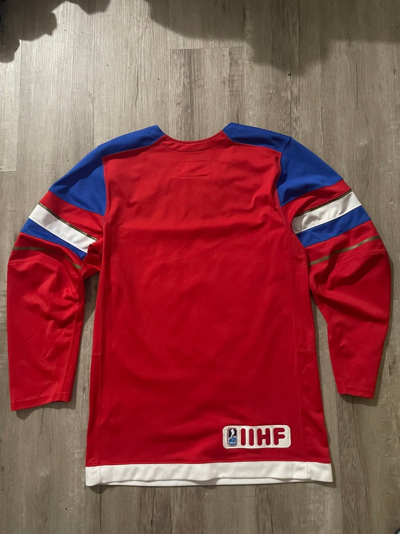 May include: Red hockey jersey with white and gold trim. The jersey has blue shoulder patches and a white patch with the IIHF logo on the back.