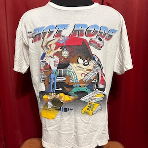 Puede incluir: Camiseta blanca con un gráfico colorido de Correcaminos y Taz, con las palabras "HOT RODS" en azul y rojo. El diseño incluye coches de dibujos animados y manuales de reparación de automóviles. Una camiseta de estilo vintage.