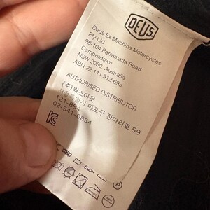 May include: A white clothing tag with the "DEUS" logo and text. The tag includes the company name, address, and authorized distributor information. Washing instructions and care symbols are also visible.