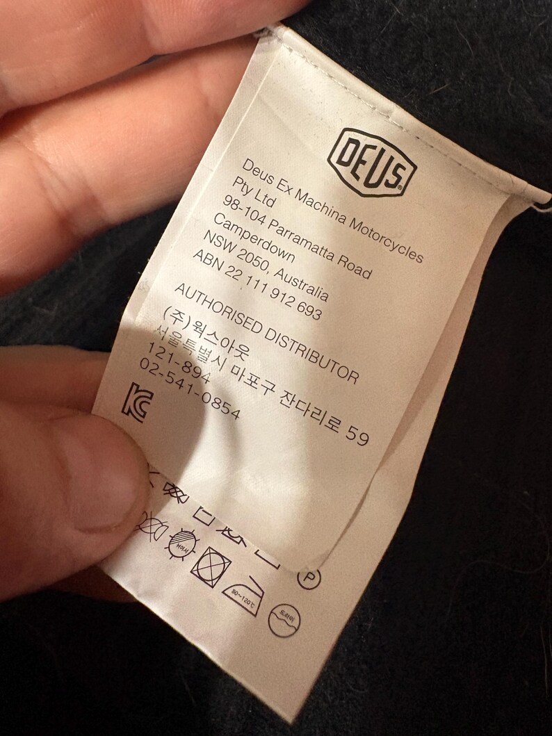 May include: A white clothing tag with the "DEUS" logo and text. The tag includes the company name, address, and authorized distributor information. Washing instructions and care symbols are also visible.
