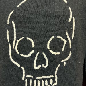 May include: Black sweater featuring a white skull design on the back. The skull is outlined with thick, textured stitching, creating a bold contrast against the dark fabric. The sweater has a classic crew neck and long sleeves.