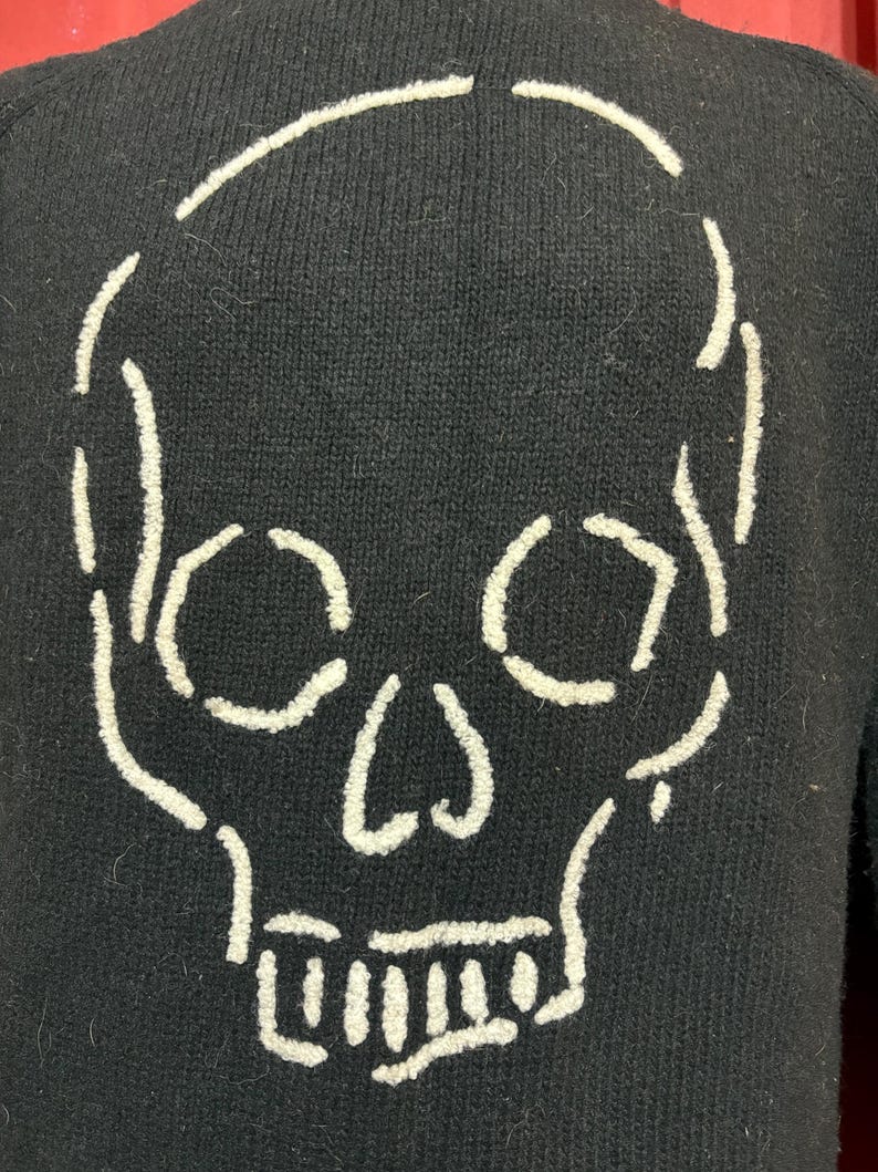 May include: Black sweater featuring a white skull design on the back. The skull is outlined with thick, textured stitching, creating a bold contrast against the dark fabric. The sweater has a classic crew neck and long sleeves.