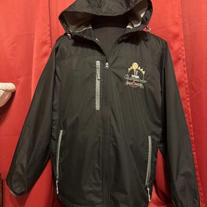 Può includere: Giacca nera con cappuccio e chiusura a zip e due tasche con zip. La giacca ha una zip grigia e dettagli grigi sulle tasche. La giacca ha un logo ricamato sul lato destro che dice "2017 NBA Champions" con una palla da basket e una rosa rossa.