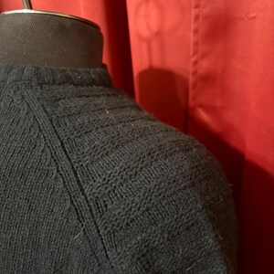 May include: Close-up of a black knit sweater with a textured pattern on the shoulder and upper sleeve. The sweater is displayed on a black mannequin head against a red backdrop.