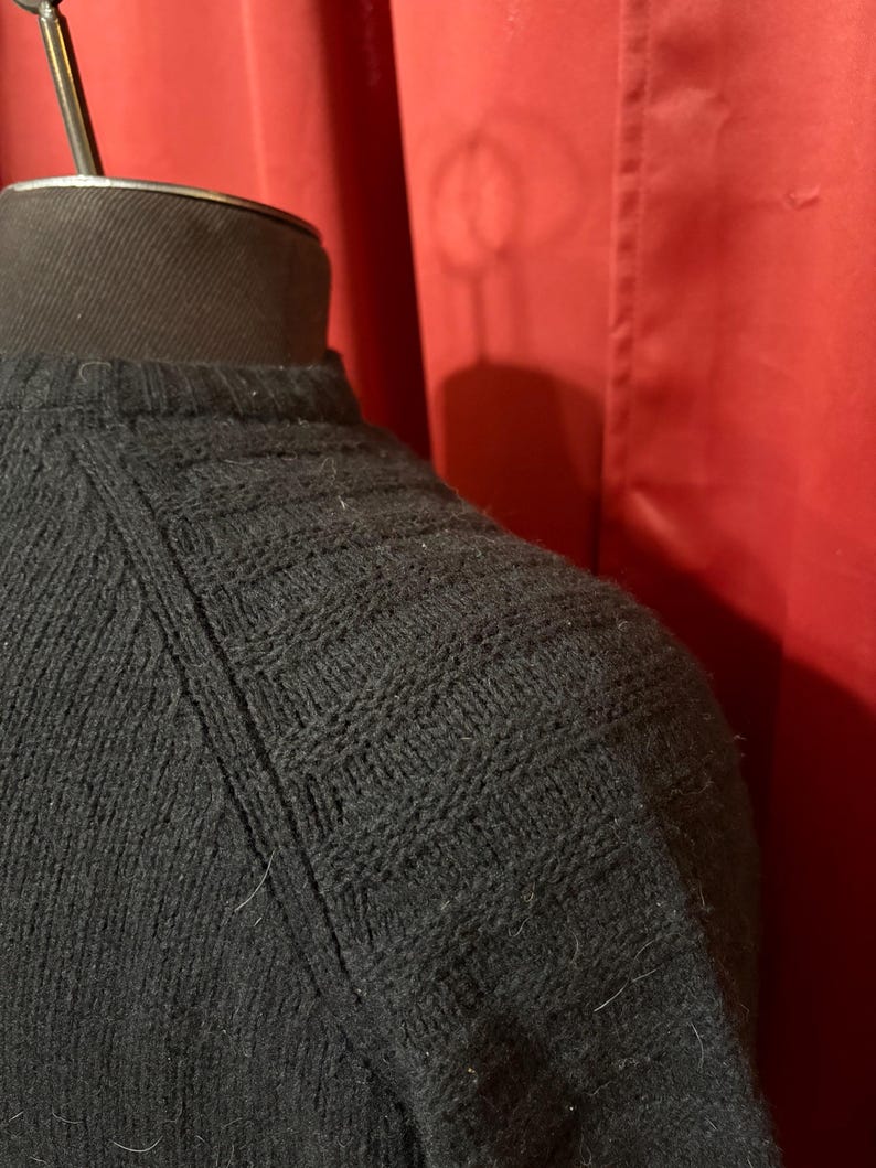 May include: Close-up of a black knit sweater with a textured pattern on the shoulder and upper sleeve. The sweater is displayed on a black mannequin head against a red backdrop.