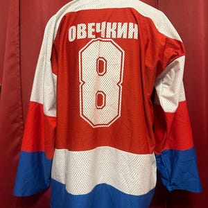 Peut inclure: Un chandail de hockey rouge, blanc et bleu avec le numéro 8 et le nom "OVЕЧКИН" au dos.
