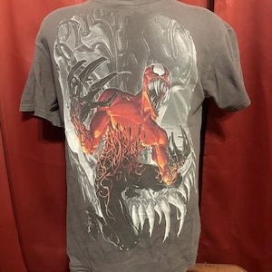 May include: Dark gray t-shirt featuring a graphic of a red and black monster with sharp claws and teeth. The design is set against a gray skull background. The shirt has short sleeves and a crew neck.
