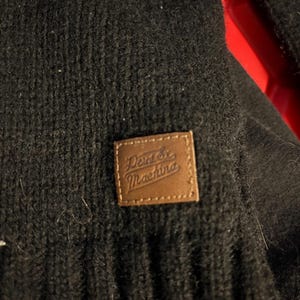 May include: Close-up of a black knit garment with a brown leather patch. The patch features the text "Deus Ex Machina" in a stylized font. The garment has a textured, ribbed appearance.