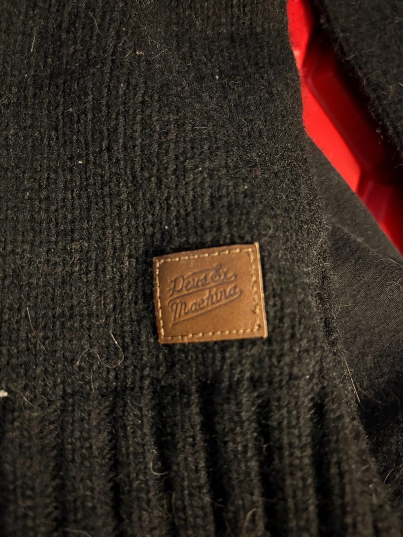 May include: Close-up of a black knit garment with a brown leather patch. The patch features the text "Deus Ex Machina" in a stylized font. The garment has a textured, ribbed appearance.