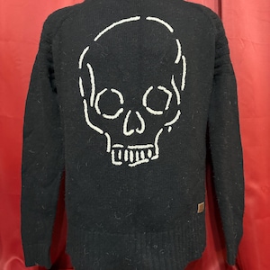 May include: Black sweater with a white skull design on the back. The skull is outlined with a thick, stitched pattern. The sweater has long sleeves and a crew neck. A small brown tag is on the lower right side.
