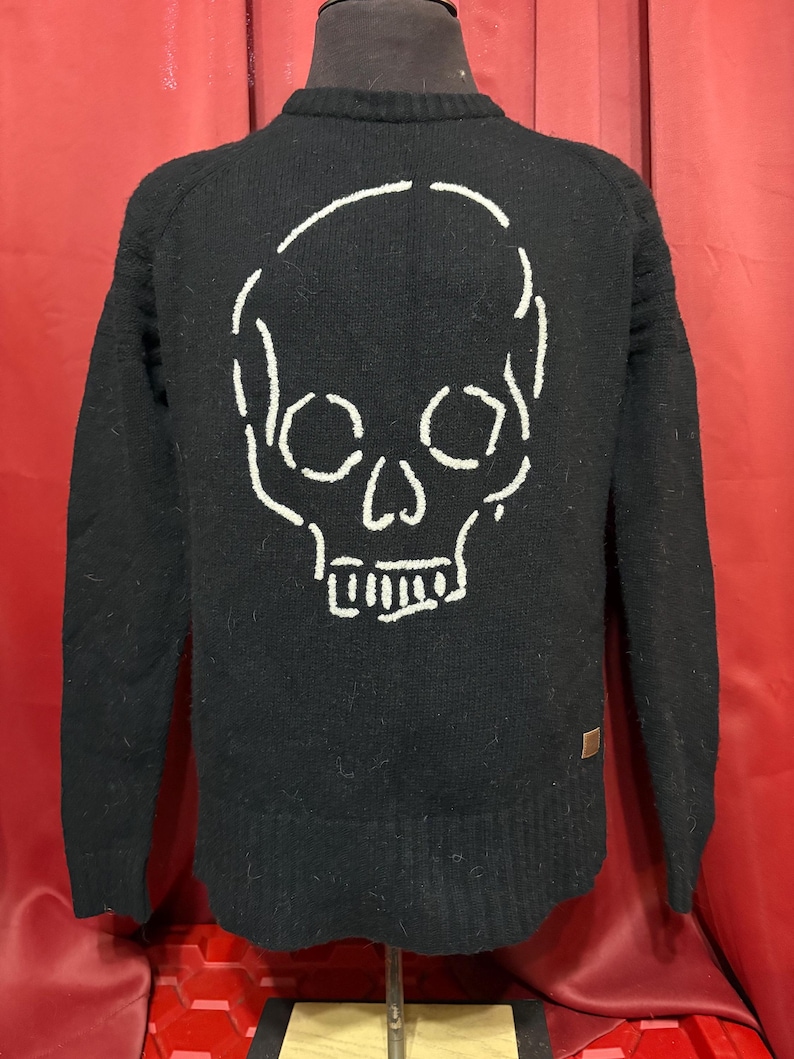 May include: Black sweater with a white skull design on the back. The skull is outlined with a thick, stitched pattern. The sweater has long sleeves and a crew neck. A small brown tag is on the lower right side.