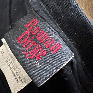 May include: Close-up of a black t-shirt tag with the words "Roman Ridge" in red gothic lettering. The tag also includes the size "XL" and the text "100% Cotton Preshrunk Made in Honduras."