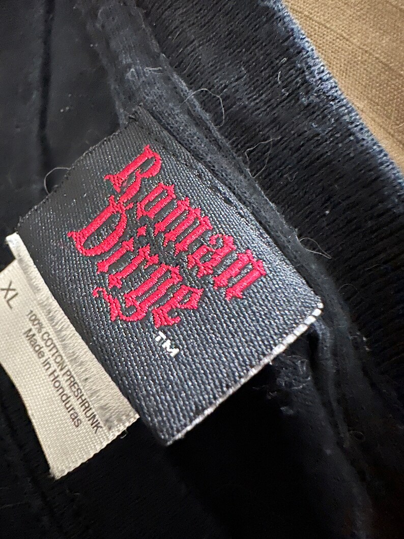 May include: Close-up of a black t-shirt tag with the words "Roman Ridge" in red gothic lettering. The tag also includes the size "XL" and the text "100% Cotton Preshrunk Made in Honduras."